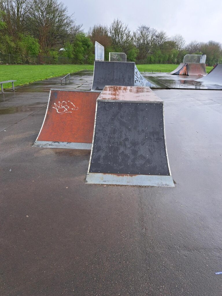 Two ramps next to each other, one with the new surfacing, the other wooden. The overview of the skatepark visible in the background.